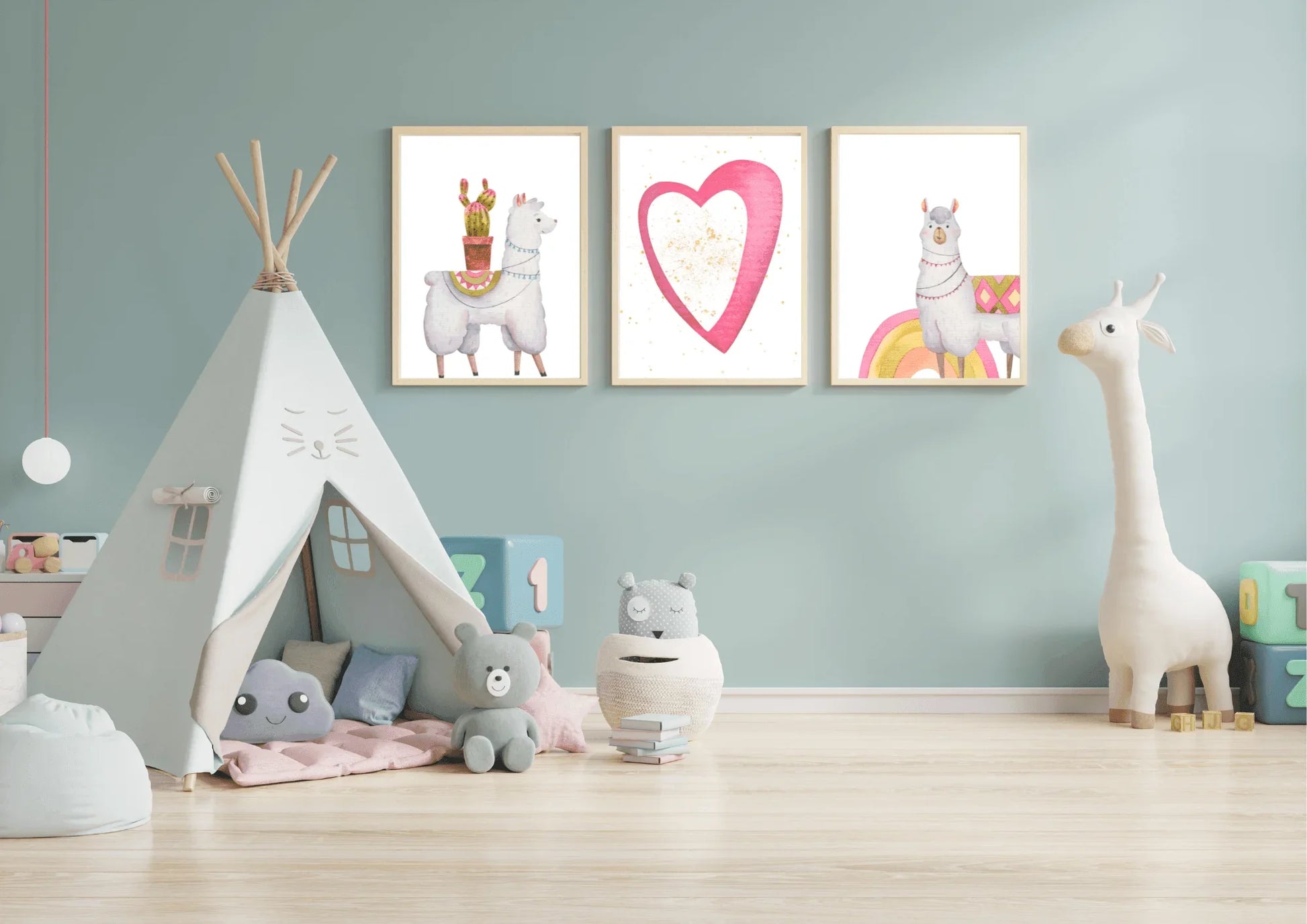 A set of three framed digital art prints featuring llamas and a heart, hanging on a light blue wall in a child's playroom. A white teepee tent, stuffed animals, and a large toy giraffe are arranged on the light wood floor.