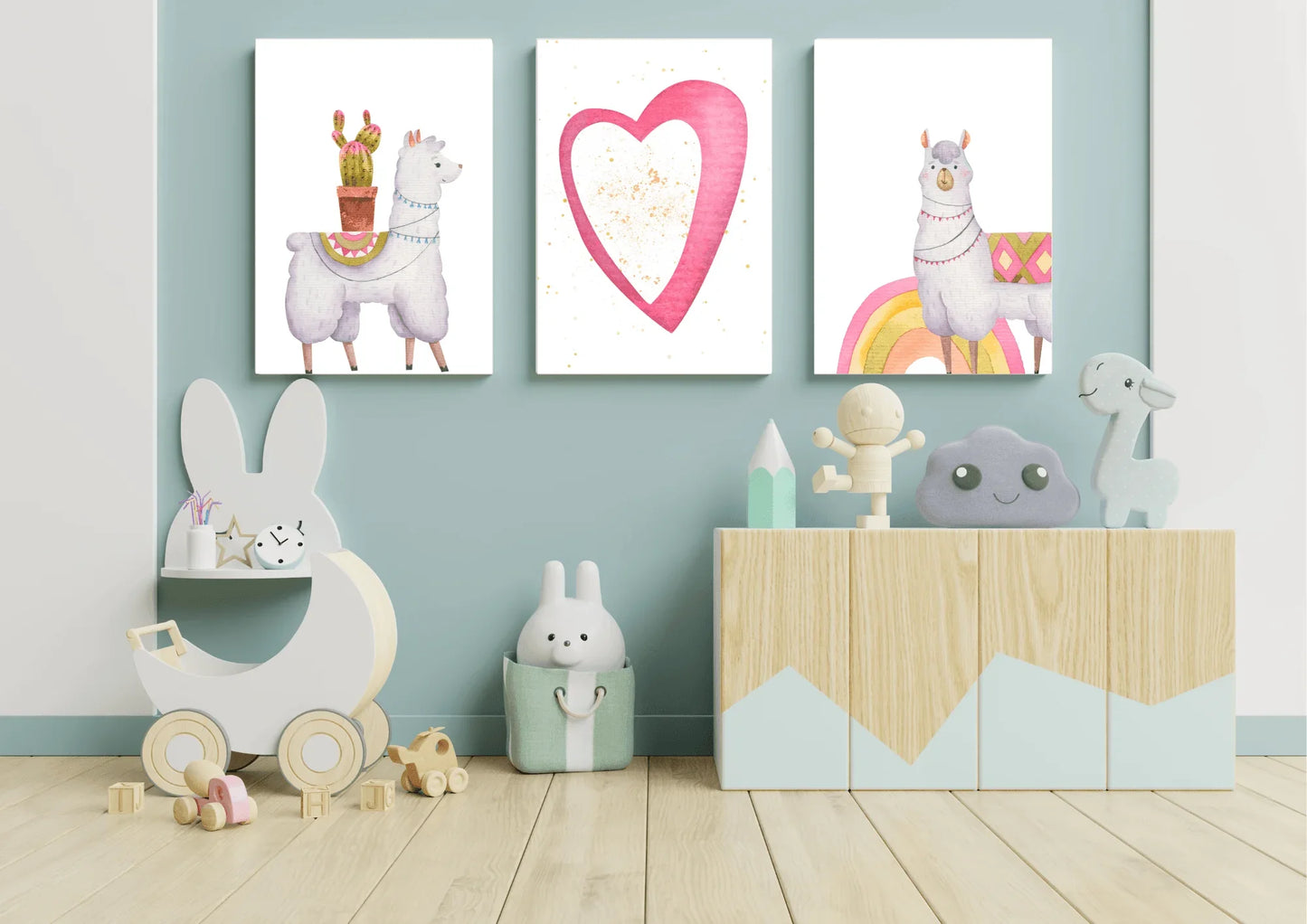 A set of three digital art prints featuring two llamas and a heart, hanging on a light blue wall. The prints are above a light wood cabinet filled with toys, and a wooden rabbit-shaped toy shelf.