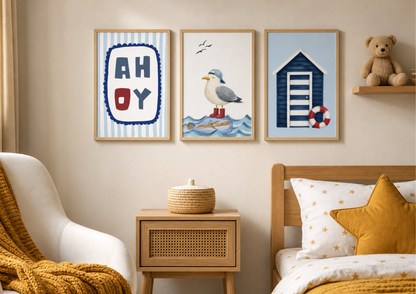 Three framed nautical-themed wall art prints hang above a bed in a styled bedroom. The prints feature a striped 'AHOY' design, a seagull on a message in a bottle, and a blue beach hut with a red life preserver.