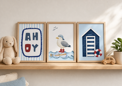 Three framed nautical-themed wall art prints displayed on a wooden shelf next to a plush bunny toy and a small potted plant. The prints feature a striped 'AHOY' sign, a seagull on a message in a bottle, and a blue beach hut with a red-and-white life preserver.