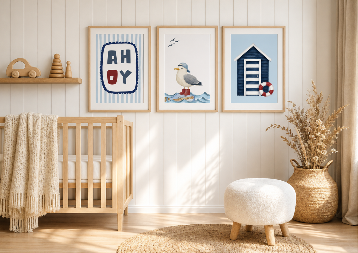 Three framed nautical-themed nursery wall art prints hang above a wooden crib in a bright, sunlit nursery. The prints feature a striped 'AH-OY' design, a seagull on waves, and a blue beach hut with a red and white life preserver.