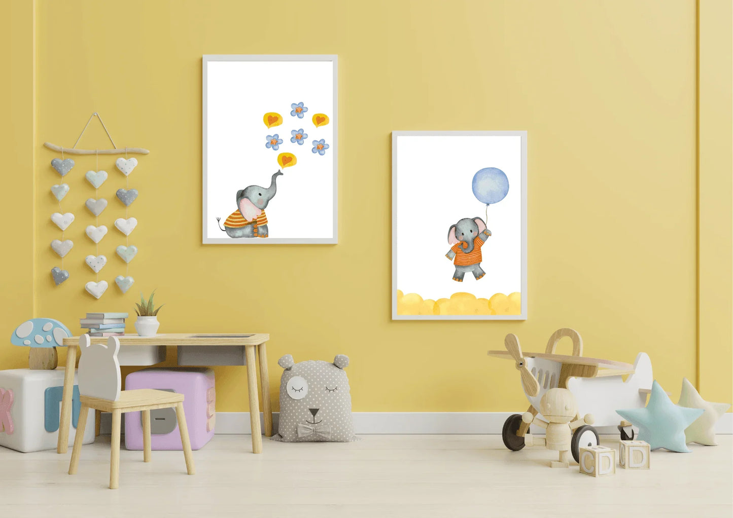 A bright yellow-walled child's room or play area featuring a small wooden desk and chair, various toys, and a heart-shaped felt wall hanging. Two framed watercolor elephant prints are displayed on the wall, one showing an elephant blowing hearts and flowers and the other flying with a blue balloon.