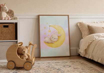 A framed pastel nursery art print featuring a cute bunny on a crescent moon, leaning against a wall in a styled children's bedroom next to a bed and wooden toy cart.
