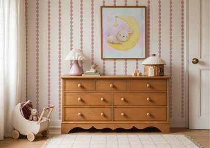A warm wood dresser in a styled nursery room with pink and white striped wallpaper, a pink lamp, a framed bunny moon print, and a toy stroller on the floor.