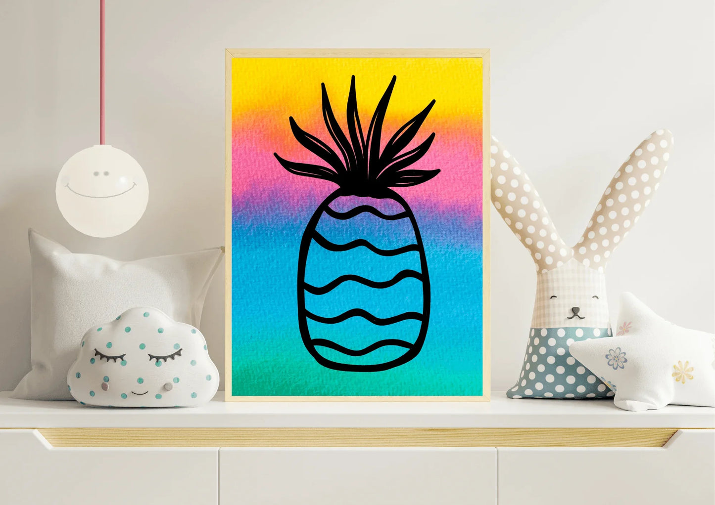 Close-up of a framed watercolor pineapple print with a bright rainbow background, sitting on a white nursery shelf next to a cloud pillow and a large stuffed bunny toy.