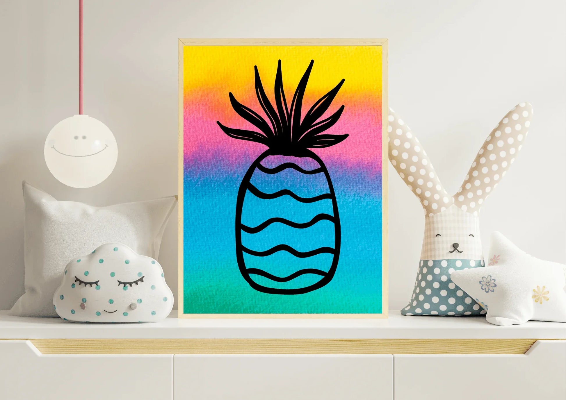 Close-up of a framed watercolor pineapple print with a bright rainbow background, sitting on a white nursery shelf next to a cloud pillow and a large stuffed bunny toy.