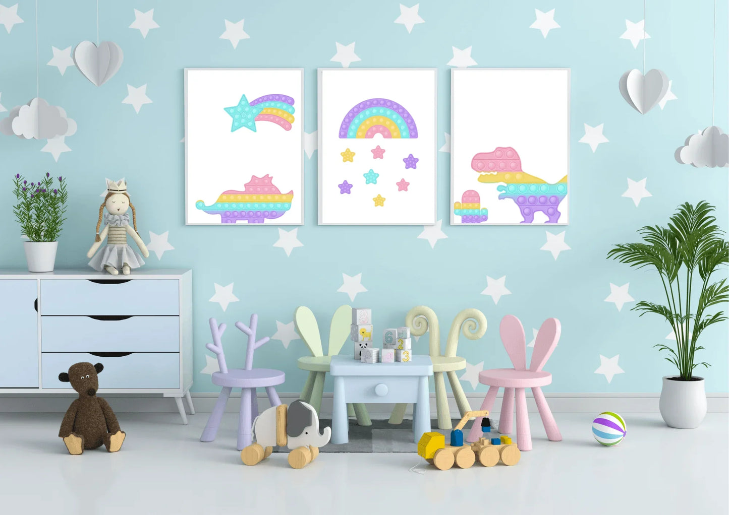 A three-piece nursery print set, featuring pop-it style dinosaurs, a rainbow, and a shooting star, hanging on a light blue wall with a white star pattern above a small table and children's toys.