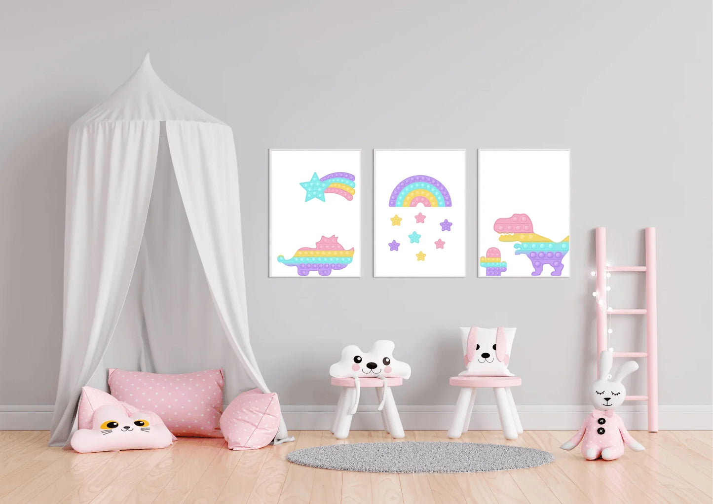 A three-piece nursery print set, featuring pop-it style dinosaurs, a rainbow, and a shooting star, hanging on a gray wall above a white bed canopy, two small chairs, and a pink ladder shelf.