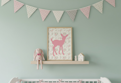 A close-up of a pink deer silhouette print framed in light wood, resting on a natural wood shelf above a white crib. The background of the print is a light floral pattern. A pink bunny plush toy and two small ceramic houses sit on the shelf. Pink and green polka-dot banner flags hang above the shelf against a soft mint green wall.