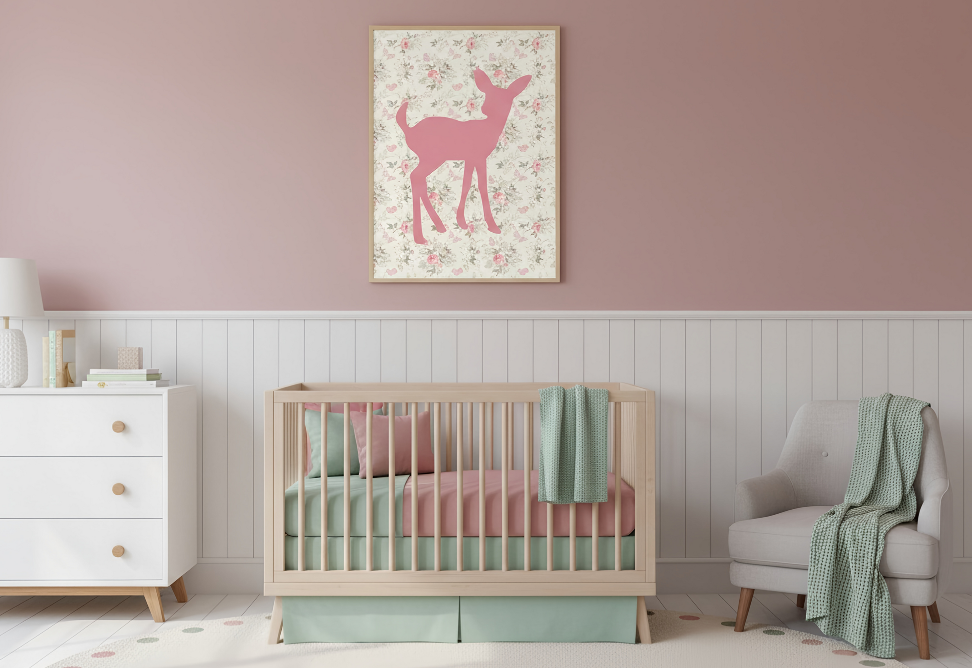 
A gender-neutral nursery features a light wood crib with pink and mint green bedding. Above the crib, a framed pink deer silhouette print on a vintage floral background hangs against a mauve pink wall, separated from white wainscoting. A white dresser is on the left, and a light gray armchair with a draped green blanket is on the right.
