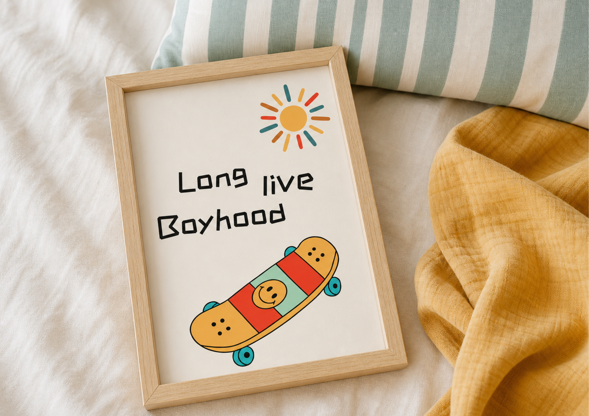 A framed art print with the text 'Long live Boyhood' and a colorful retro sun and skateboard illustration, styled on a white textured blanket with a striped pillow and yellow fabric in the background.