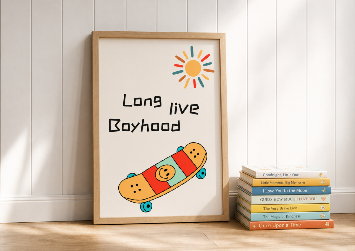 A framed art print leaning against a white paneled wall on a light wood floor, next to a stack of colorful books. The print features the text 'Long live Boyhood' with a hand-drawn skateboard and sun illustration.