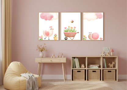 A set of three charming watercolor nursery prints displayed over a small wooden desk and cube shelf in a child's bedroom with pink walls. The prints feature a pink chick, a wheelbarrow filled with flowers and a bee, and a pink snail, all beneath pink clouds.