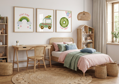 A brightly lit child's bedroom with a gallery wall featuring three framed watercolor prints: a rainbow, a carrot truck, and a green donut. A rattan bed with pink and green bedding is on the right, next to a wooden bookshelf and large window. The room includes a wooden desk, rattan chair, round jute rug, and several woven baskets.