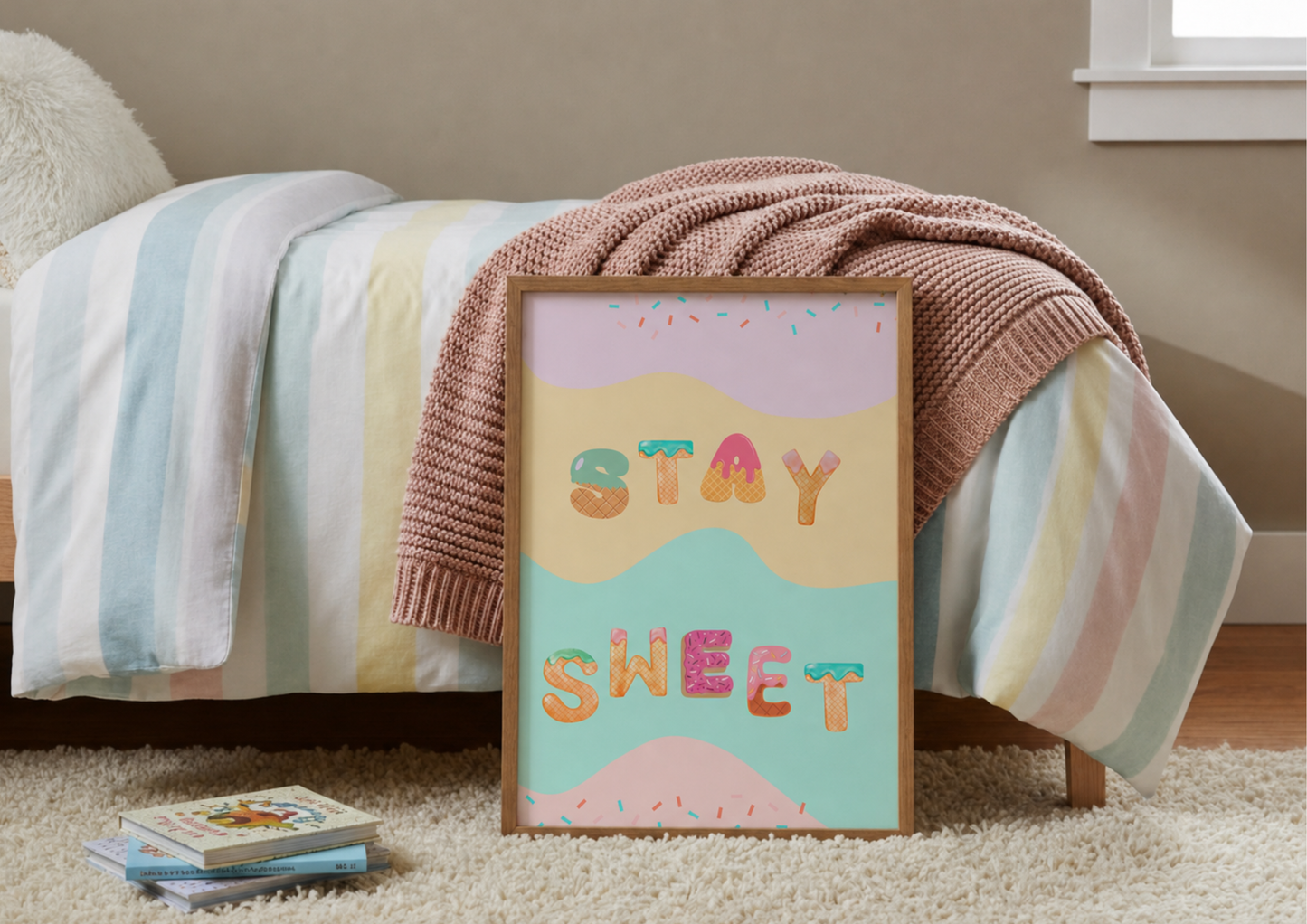A framed pastel art print leaning against a bed in a bedroom. The print says “STAY SWEET” in colorful letters over pink, cream, and mint sections, with a pink knit blanket draped over the bed and striped bedding in the background.