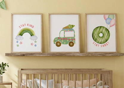 A close-up shot of three framed art prints displayed side-by-side on a rustic wood shelf above a natural wood crib against a pale yellow wall. The prints are watercolors: the first is a pink and green rainbow with "stay kind," the second is a green truck carrying a large carrot, and the third is a green donut with balloons and "stay sweet."