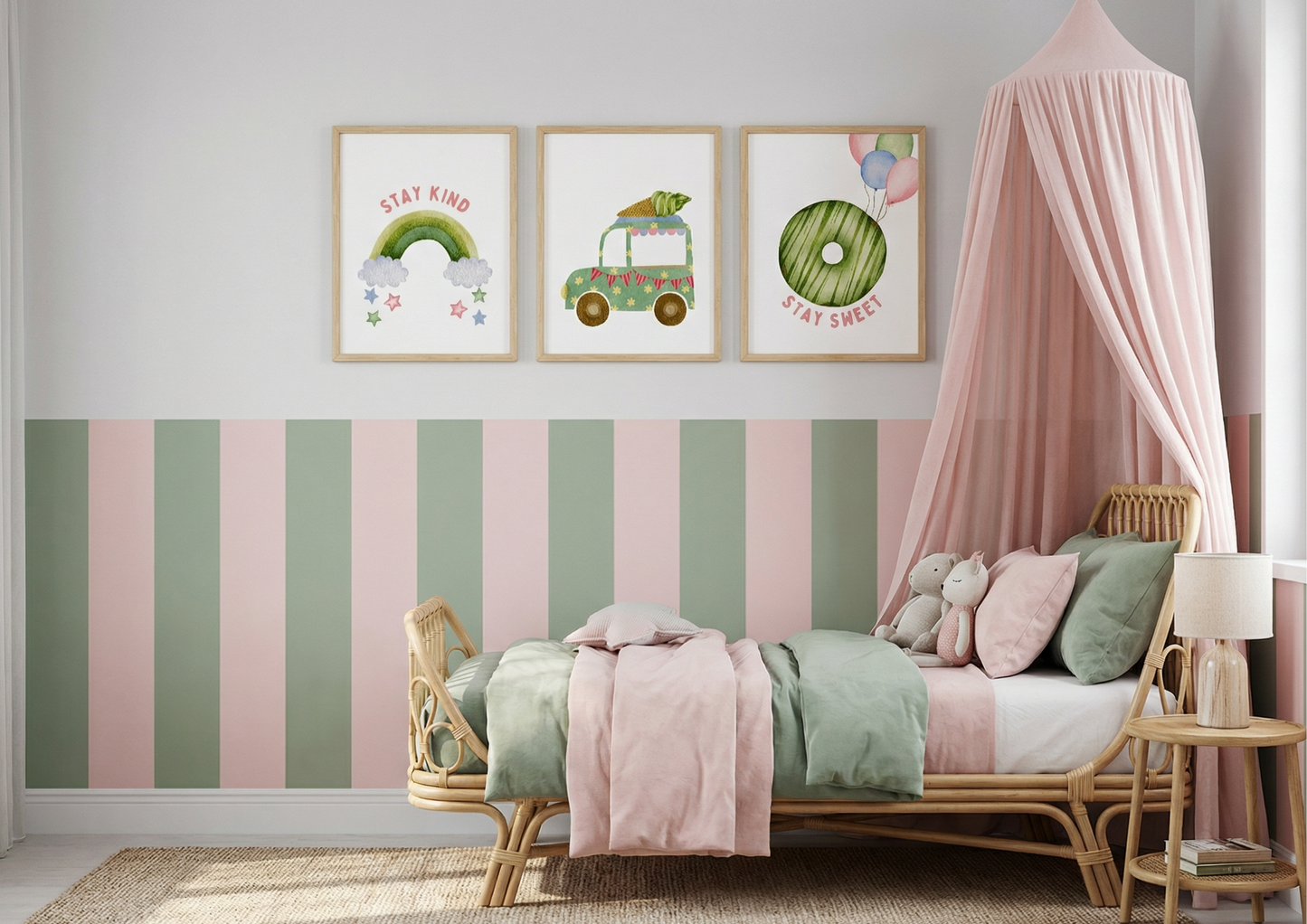 Three framed watercolor art prints hang above a child's rattan bed against a wall painted with horizontal pink and olive green stripes on the bottom half. The prints feature, from left to right: a pink and green rainbow with "stay kind," a green carrot truck, and a green donut with balloons and "stay sweet." The bed is dressed in pink and mint green bedding, and a pink canopy drapes over the right side.