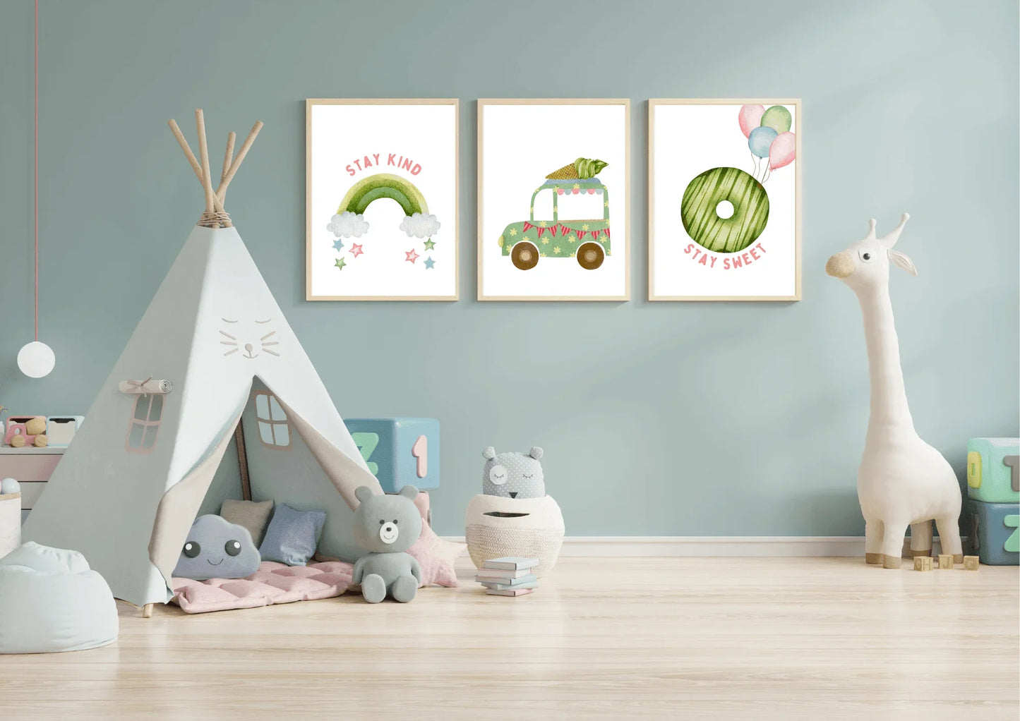 Three framed watercolor art prints hang above a teepee on a light blue wall in a child's playroom. The prints feature, from left to right: a pink and green rainbow with "stay kind," a green truck carrying a large carrot, and a green donut with balloons and "stay sweet." The room also has a large white stuffed giraffe and various soft toys.