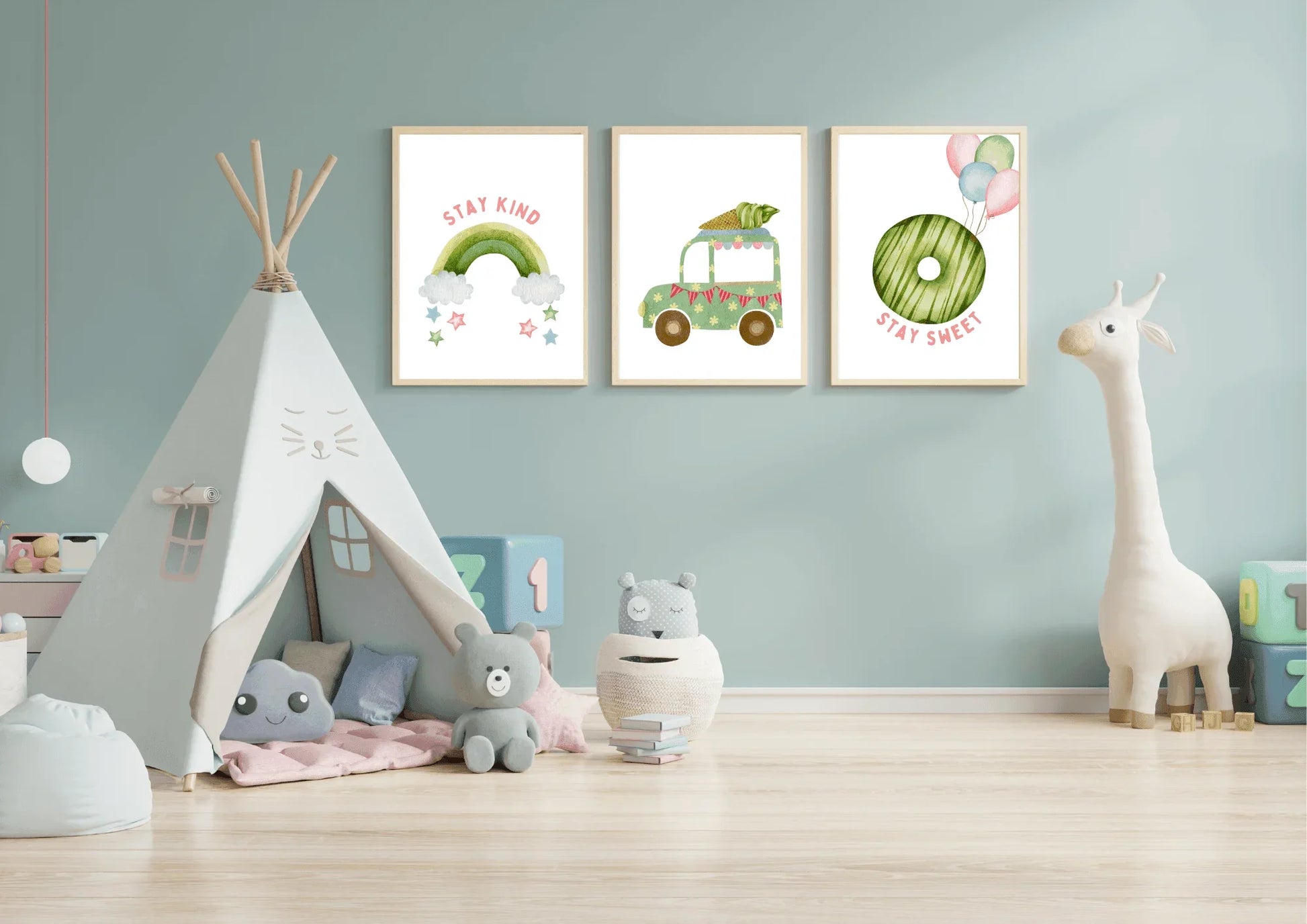 Three framed watercolor art prints hang above a teepee on a light blue wall in a child's playroom. The prints feature, from left to right: a pink and green rainbow with "stay kind," a green truck carrying a large carrot, and a green donut with balloons and "stay sweet." The room also has a large white stuffed giraffe and various soft toys.