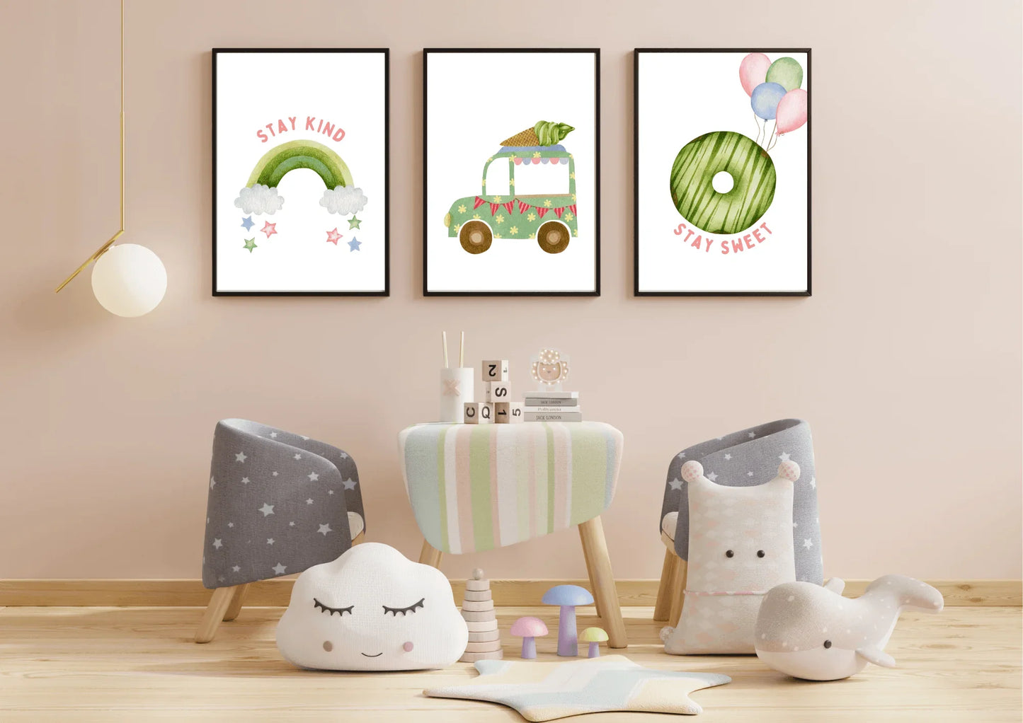 Three framed watercolor art prints with black frames hang above a small, light wood play table against a pale pink wall. The prints feature, from left to right: a green rainbow with "stay kind," a green ice cream truck, and a green donut with balloons and "stay sweet." The play space below has two small chairs covered in gray star fabric, a cloud pillow, and soft animal toys.