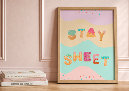 A framed pastel art print leaning against a pink paneled wall on a wooden floor, next to a stack of books. The print features the words 'STAY SWEET' in decorative icing-like letters over wavy pastel shapes.