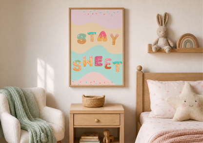 A framed nursery wall art print with the words 'STAY SWEET' in colorful letters hangs above a wooden nightstand in a softly styled children's bedroom. The print has pastel wavy shapes and confetti details, with a bunny on a shelf and a star pillow nearby.