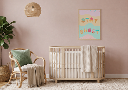 A styled nursery interior with a light wood crib, rattan chair, hanging pendant light, plant, and a framed pastel wall print reading “STAY SWEET.”.