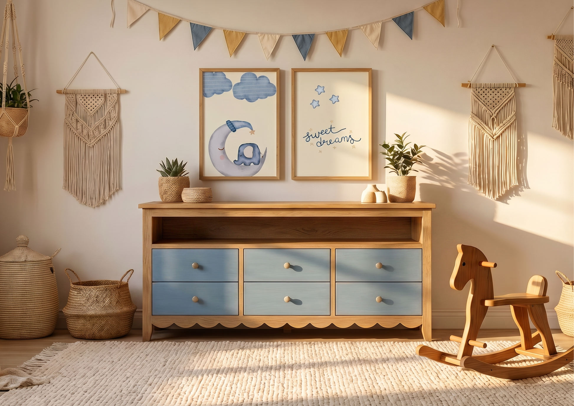 A cozy, sunlit nursery featuring a light wood dresser with six soft blue drawers and a scalloped bottom edge. Above the dresser, two wooden-framed art prints are displayed: one of a whimsical elephant sleeping on a crescent moon and another with the text "sweet dreams" surrounded by stars. The wall is decorated with a blue, cream, and mustard fabric bunting and two macrame wall hangings. A wooden rocking horse sits on a cream textured rug in the foreground, with woven baskets and small potted plants adding 
