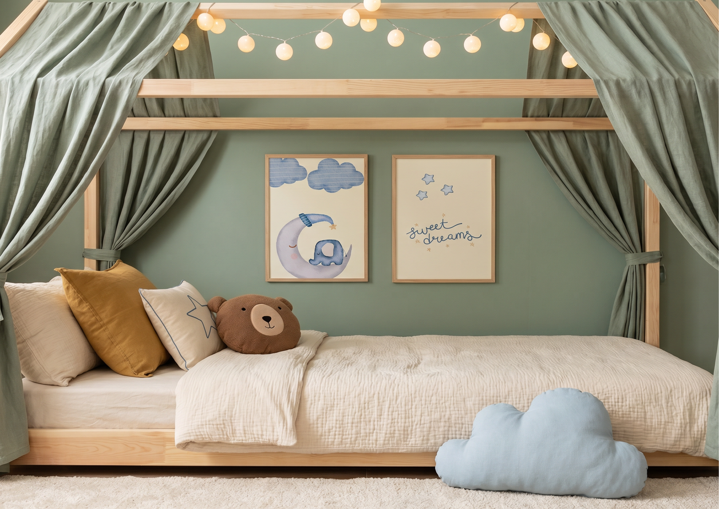 A peaceful, nature-themed Montessori floor bed with a light wood house frame and draped sage green linen curtains. The bed is styled with a cream textured duvet, a mustard yellow pillow, a brown bear-face cushion, and a soft blue cloud pillow on the rug nearby. On the sage green wall behind the bed, two wooden-framed prints are displayed: one featuring a whimsical elephant on a crescent moon and the other with "sweet dreams" in blue script. A string of warm, glowing globe lights is draped across the top of 