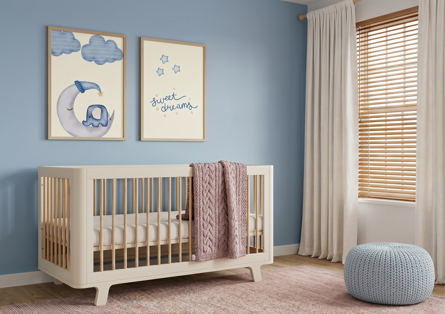 A modern and calming nursery featuring a soft blue accent wall. In the center, a cream-colored crib with light wood spindles is styled with a dusty pink cable-knit blanket. Above the crib, two wooden-framed art prints are displayed: one of a whimsical elephant sleeping on a crescent moon and another with the text "sweet dreams" in blue script. To the right, a window with wooden blinds and floor-to-ceiling cream curtains lets in gentle light, while a light blue textured floor pouf sits on a pinkish-toned rug