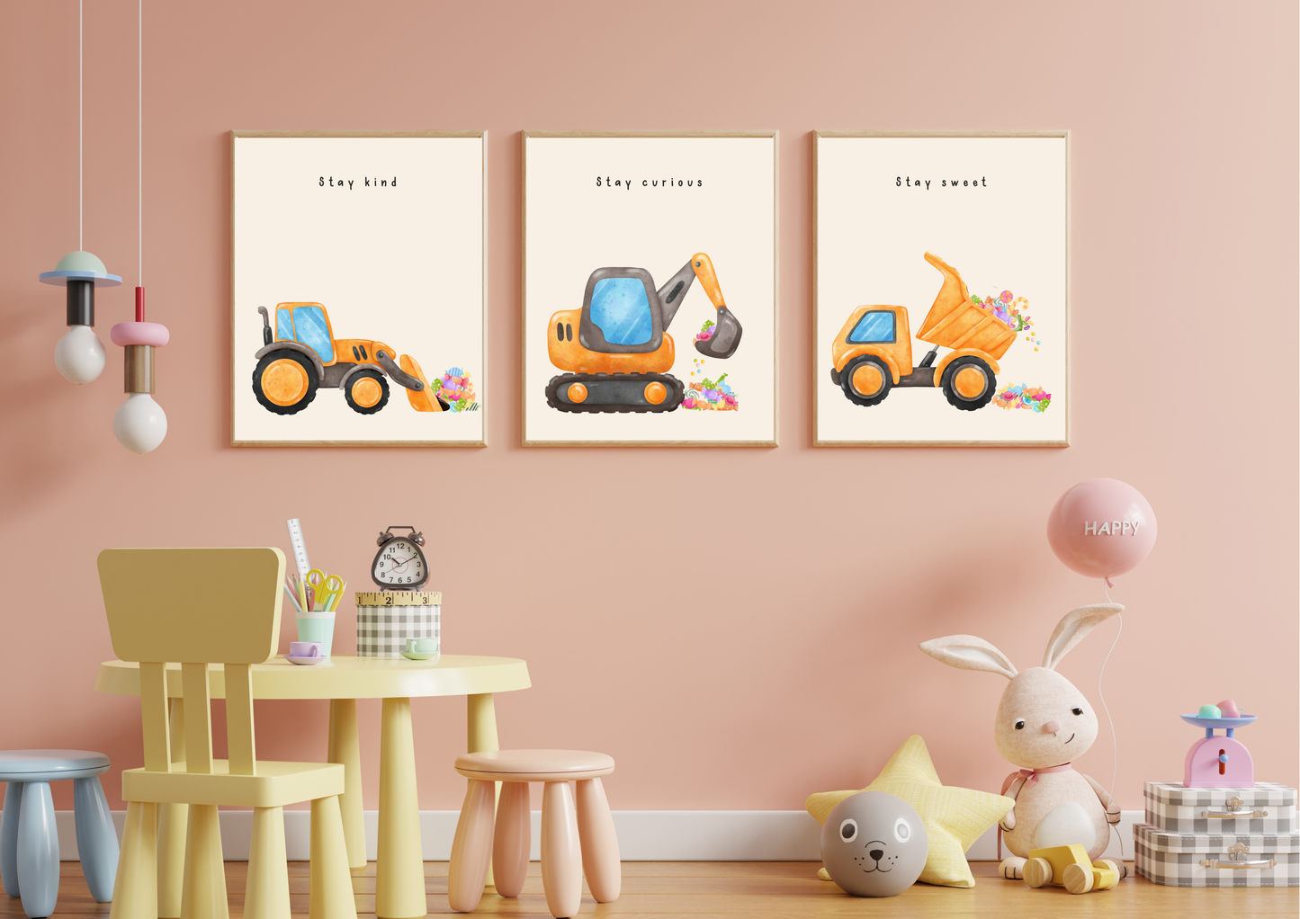 Set of three framed construction vehicle prints (Stay Kind, Stay Curious, Stay Sweet) hanging above a small yellow table and chairs in a pink-walled playroom.