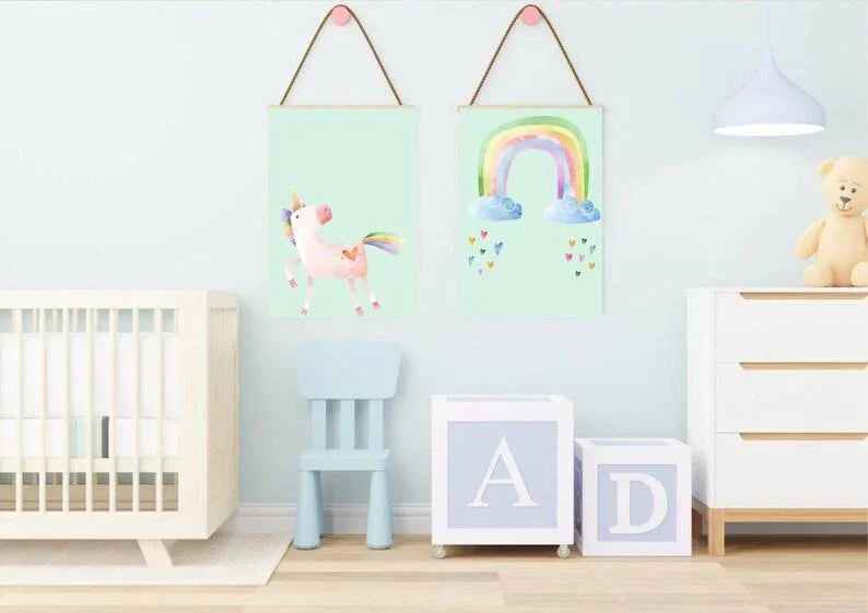 Two unicorn and rainbow wall art prints with wooden hangers on a light blue wall in a nursery with a crib and large letter blocks.