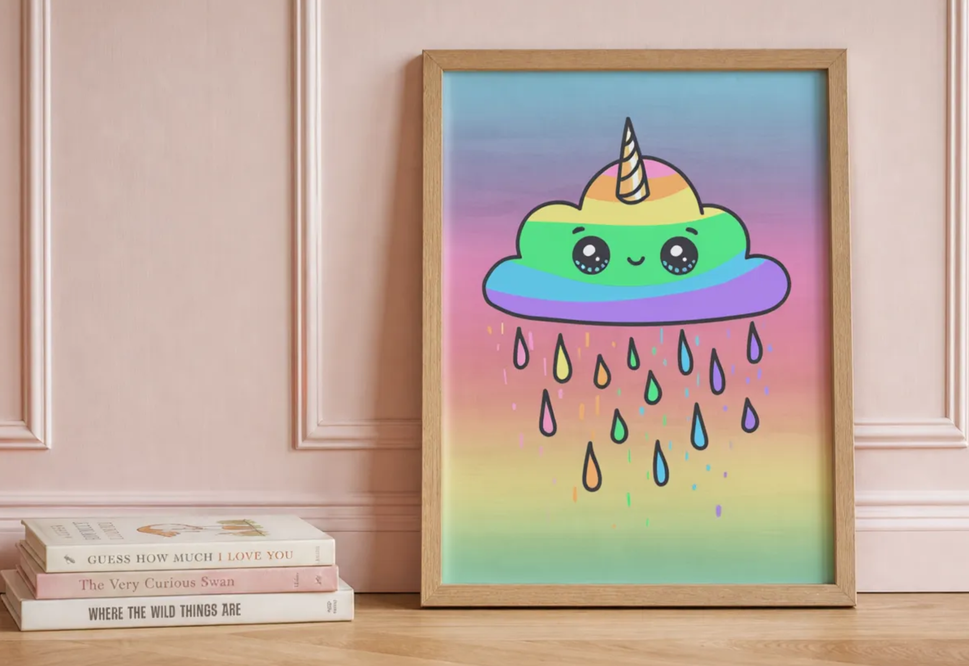 A framed pastel art print featuring a cute rainbow-colored cloud character with a horn and raindrops, leaning against a pink paneled wall on a wooden surface next to a stack of books.