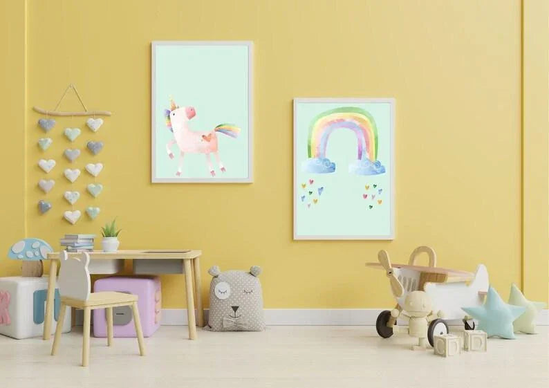 Two framed digital nursery art prints featuring a watercolor unicorn and a rainbow, hanging on a yellow wall in a child's playroom.