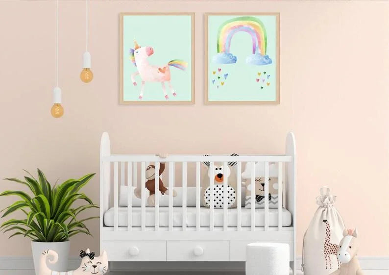 Two framed digital art prints of a unicorn and a rainbow with wooden frames, hanging on a light pink wall above a white crib.