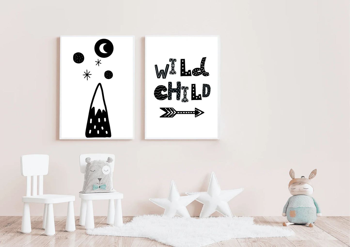 Mockup of two gender-neutral black and white wall art posters in a soft pink nursery. The prints feature a stylized mountain design and the quote "WILD CHILD". The foreground includes a white sheepskin rug, two white star pillows, and a children's toy.