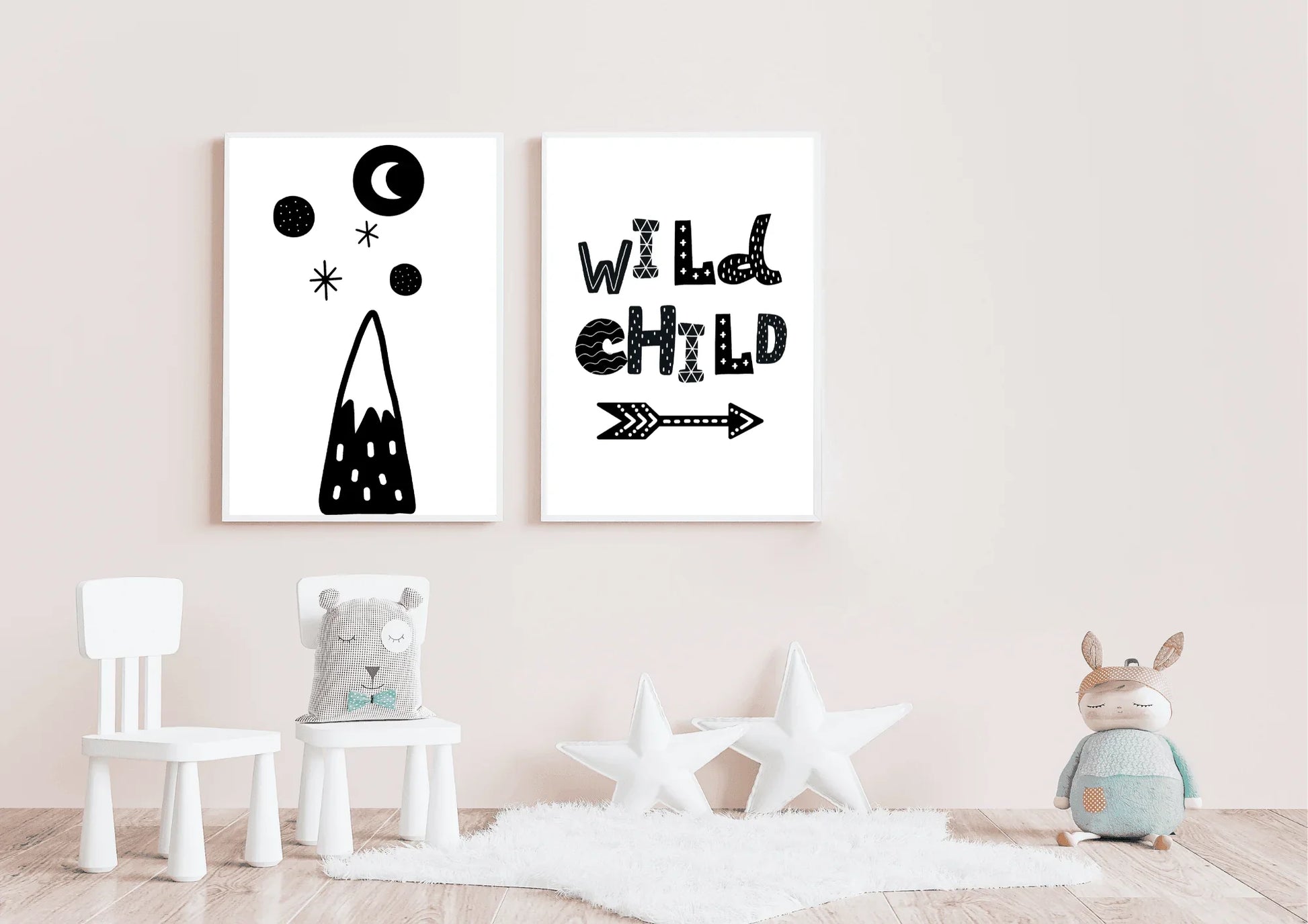Mockup of two gender-neutral black and white wall art posters in a soft pink nursery. The prints feature a stylized mountain design and the quote "WILD CHILD". The foreground includes a white sheepskin rug, two white star pillows, and a children's toy.
