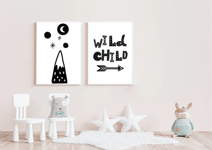Mockup of two gender-neutral black and white wall art posters in a soft pink nursery. The prints feature a stylized mountain design and the quote "WILD CHILD". The foreground includes a white sheepskin rug, two white star pillows, and a children's toy.