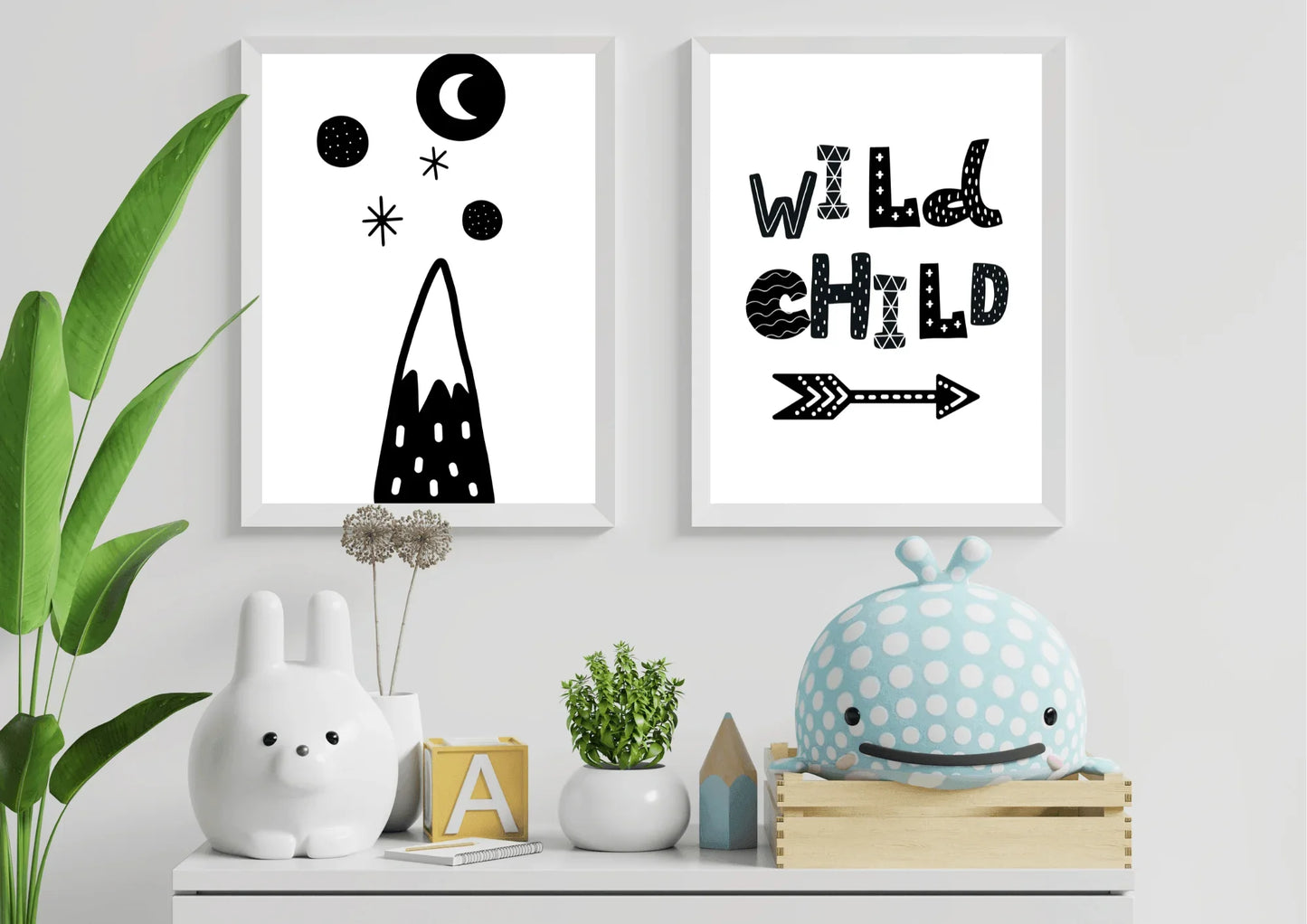 Two white-framed black and white nursery art prints on a dresser, one with a stylized mountain, moon, and stars, and the other reading "WILD CHILD" with an arrow. The prints are surrounded by a white rabbit sculpture, a blue polka-dot whale toy, and small potted plants.