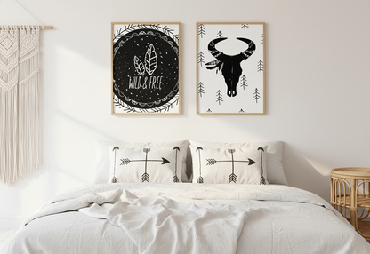 A serene bohemian-style bedroom featuring two wooden-framed art prints centered above a bed with white linens and arrow-patterned pillows. The left print shows a "Wild & Free" design with feathers on a black chalkboard-style background, while the right print features a black bull skull with a feathered headdress on a white background with small pine tree motifs. A large macramé wall hanging is visible on the left, and a small rattan side table sits to the right, all bathed in soft, natural morning light.
