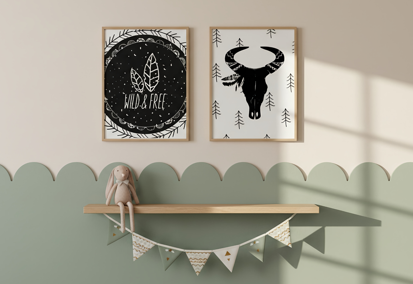 A peaceful nursery wall with a two-tone design featuring a cream upper half and a sage green lower half with a scalloped edge. Centered on the cream section are two wooden-framed art prints: one with a "Wild & Free" feather design on a black background and the other featuring a black bull skull with a feathered headdress on a white background with pine trees. Below the art, a light wood floating shelf holds a soft plush bunny, while a patterned bunting in green, cream, and gold hangs gracefully beneath the 