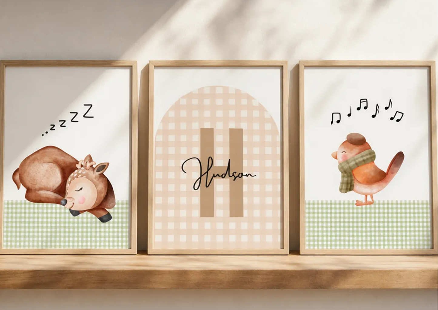 Three framed nursery art prints displayed side-by-side on a wooden shelf. The left print shows a sleeping deer with 'zzz' text, the middle print has a beige gingham arch with the name 'Hudson' in script, and the right print shows a singing bird with musical notes.