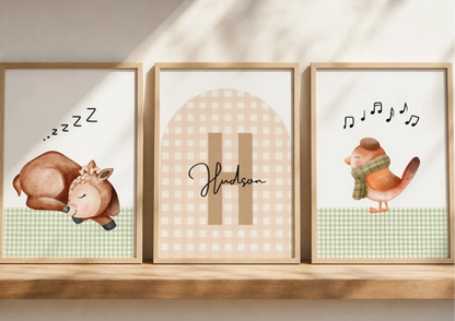 Three framed nursery art prints displayed side-by-side on a wooden shelf. The left print shows a sleeping deer with 'zzz' text, the middle print has a beige gingham arch with the name 'Hudson' in script, and the right print shows a singing bird with musical notes.
