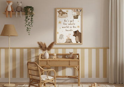 
A cozy nursery or child's room corner featuring a rattan desk and chair. Hanging above the desk is a framed Christian wall art print with the lyric "He's got the whole world in His hands" surrounded by cute woodland animals in a soft, neutral color palette. The lower half of the wall is decorated with wide vertical stripes in cream and light yellow.
