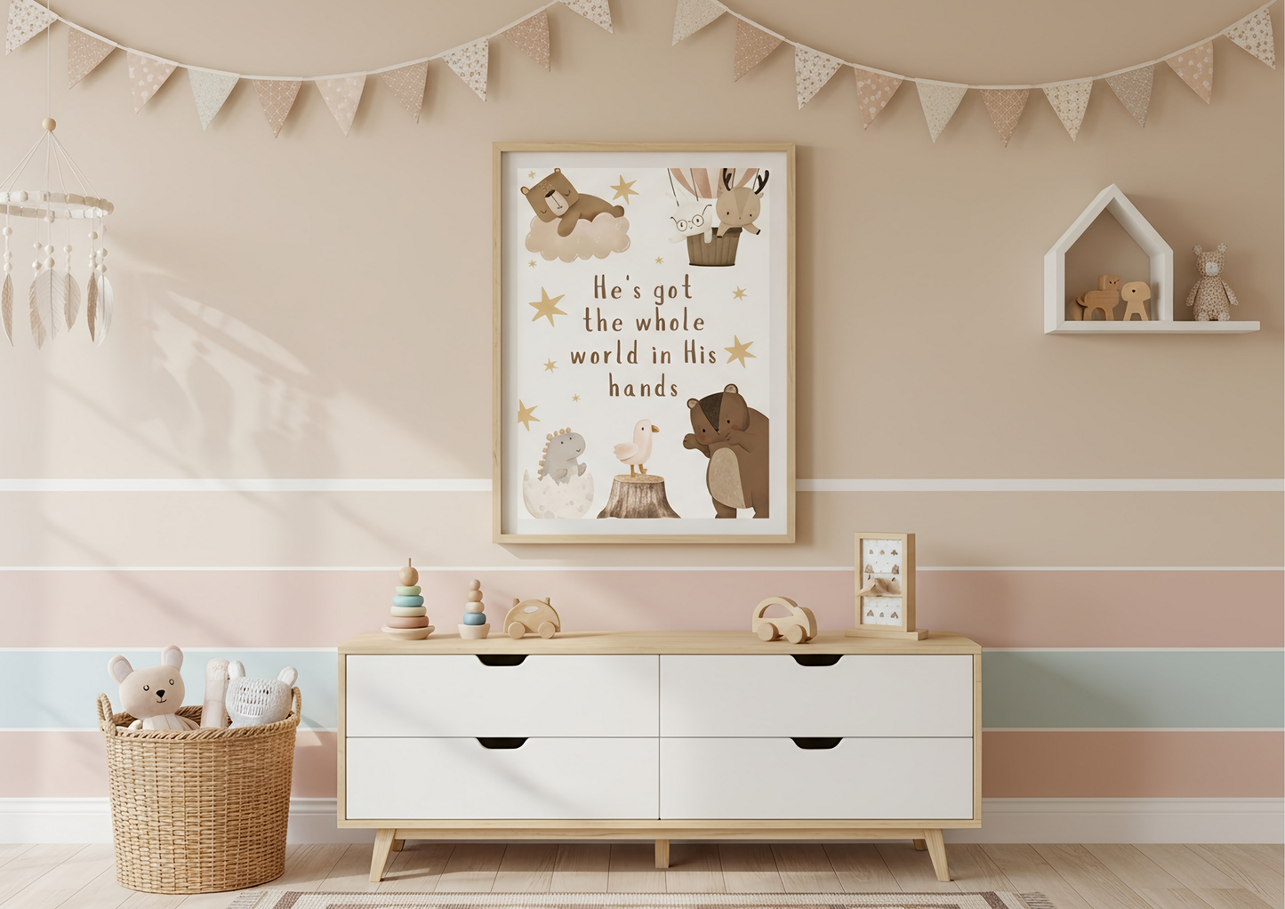 A nursery scene featuring a framed Christian wall art print with the lyric "He's got the whole world in His hands" and cute woodland animals, hanging above a white mid-century modern dresser. The wall is decorated with horizontal stripes in soft beige, pink, and pale mint green. A neutral-toned bunting hangs near the ceiling, and a house-shaped shelf with small toys is on the right.