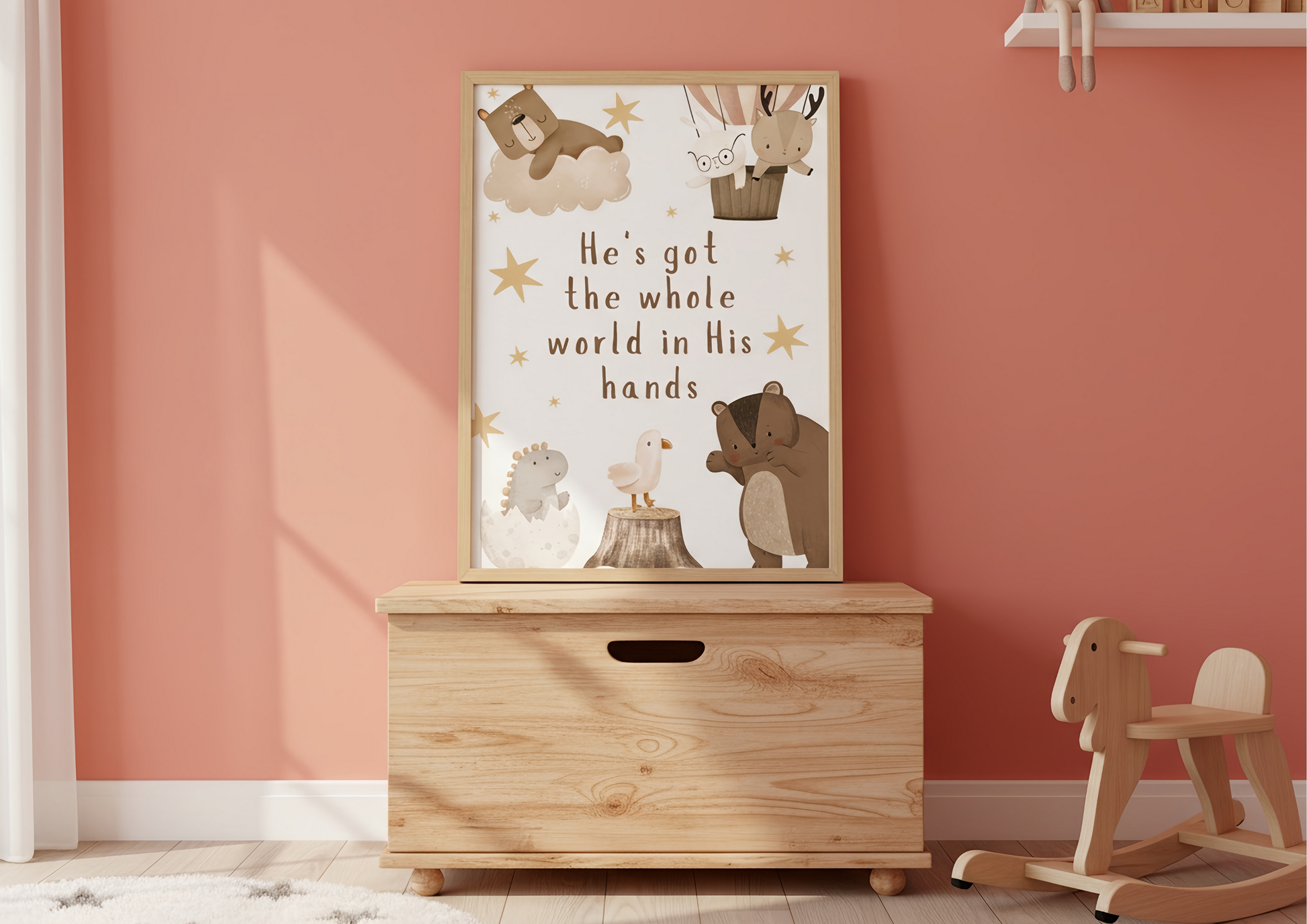 
A close-up of a child's room featuring a framed Christian woodland art print with the phrase "He's got the whole world in His hands" and cute forest animals, leaning against a coral pink wall. The print rests on a light wood toy storage chest next to a wooden rocking horse.