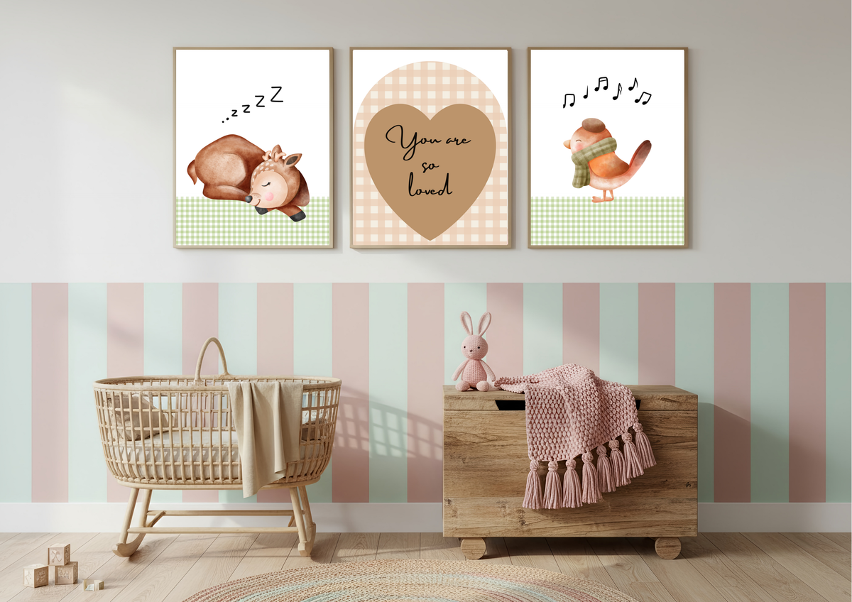 Three framed nursery wall art prints hang above a wooden toy chest and wicker bassinet in a softly styled nursery room. The prints feature a sleeping deer with 'zzz', a central heart reading 'You are so loved', and a singing bird with musical notes.