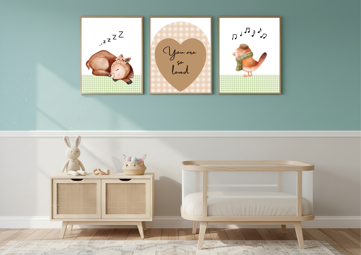 Three framed nursery wall art prints hang above a light wood cabinet and a wooden baby crib in a styled nursery room. The prints feature a sleeping baby goat, a heart with the words 'You are so loved', and a singing bird.