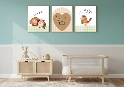 Three framed nursery wall art prints hang above a light wood cabinet and a wooden baby crib in a styled nursery room. The prints feature a sleeping baby goat, a heart with the words 'You are so loved', and a singing bird.
