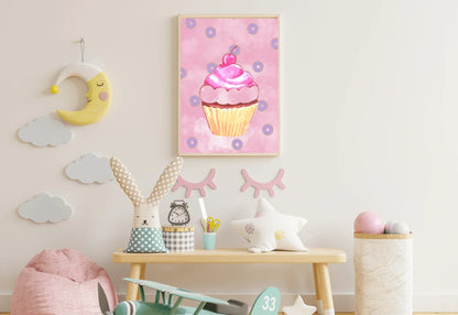 A photorealistic photograph of a bright, modern nursery with cream-colored walls and light wood flooring. The focal point is a natural wood play table featuring a whimsical rabbit plush toy with polka dots, a small checkered storage tin, and a star-shaped pillow. Hanging centrally above the table is a light wood-framed vertical art print of a pink watercolor cupcake with purple donuts. The wall is decorated with a yellow crescent moon hanging and several grey cloud decals.